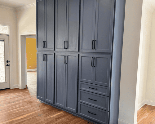 Tall, slate-blue built-in cabinetry with shaker-style doors, drawers, and black handles against a white wall and wood floor.