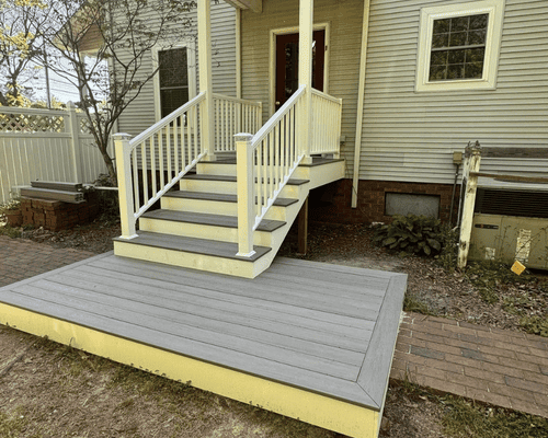 A wide wooden deck with matching stairs leads up to the front entrance of a house with yellow siding and white railings.