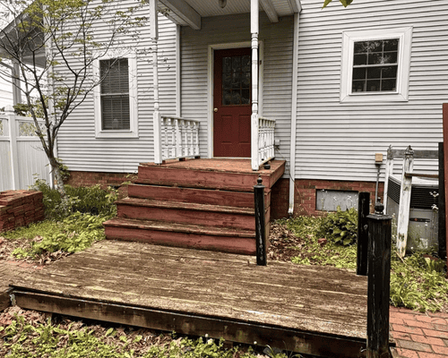 A front entrance to a house with wooden steps leading to a red door, surrounded by a worn deck and overgrown garden.