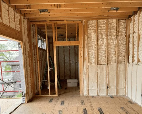 An interior view of a room under construction, featuring exposed wooden framing and walls insulated with spray foam.