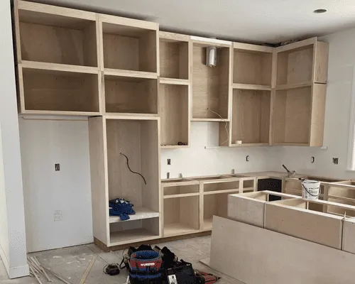 Unfinished light wood kitchen cabinets installed in a home undergoing renovation, with tools on the floor.