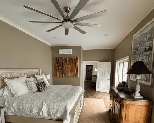 A neutral-toned bedroom features a large bed with white bedding, a modern ceiling fan, wall art, and a wooden dresser.
