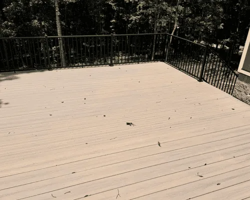 A light-colored composite deck with a black metal railing overlooking a wooded area, scattered with a few fallen leaves.