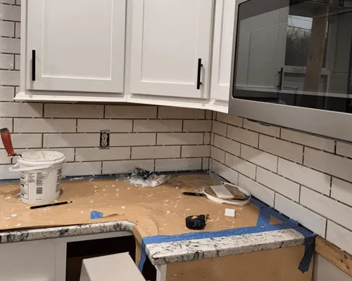A kitchen under renovation showing white subway tile backsplashes, white cabinets, and a granite countertop partially taped.