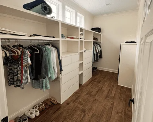 A walk-in closet featuring white wooden shelving, a built-in dresser, hanging clothes, and wood-look flooring.