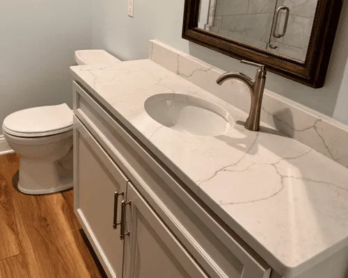 Bathroom vanity with white marble-patterned countertop, gold faucet, and white cabinet beside a toilet on wood flooring.