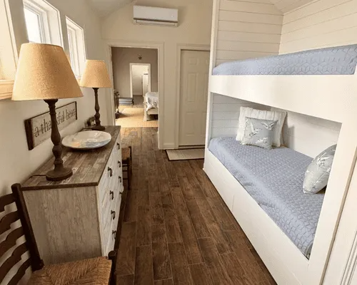 A rustic bedroom with wood flooring, built-in bunk beds with blue bedding, and a white dresser with two lamps.