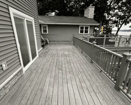 A wide, elevated grey wooden deck with a matching railing attached to the side of a grey house with a stone foundation.