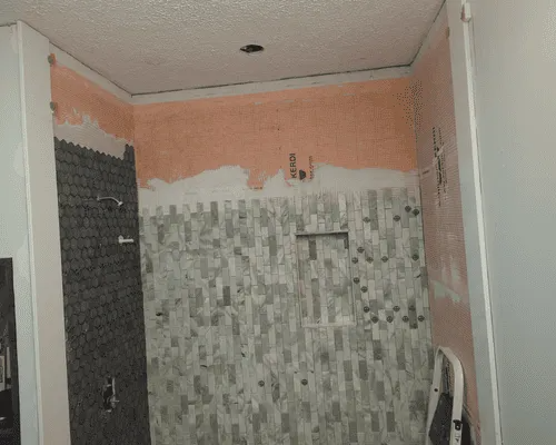 A partially tiled shower enclosure with gray patterned tiles, an open niche, and unfinished walls with exposed drywall.