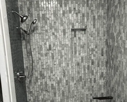 A shower with light gray marble vertical tile, a showerhead with a handheld sprayer, and a built-in recessed wall niche.