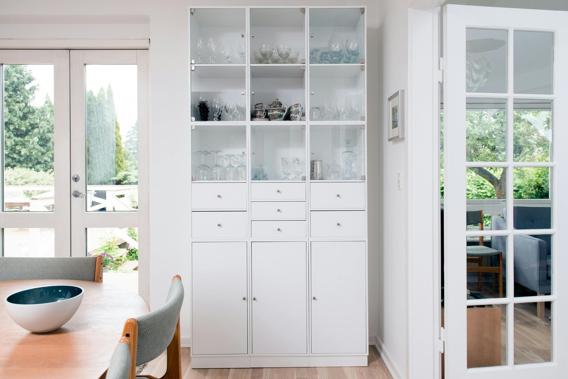 A white dining room hutch with glass-fronted upper cabinets sits between a French door and a wooden dining table.