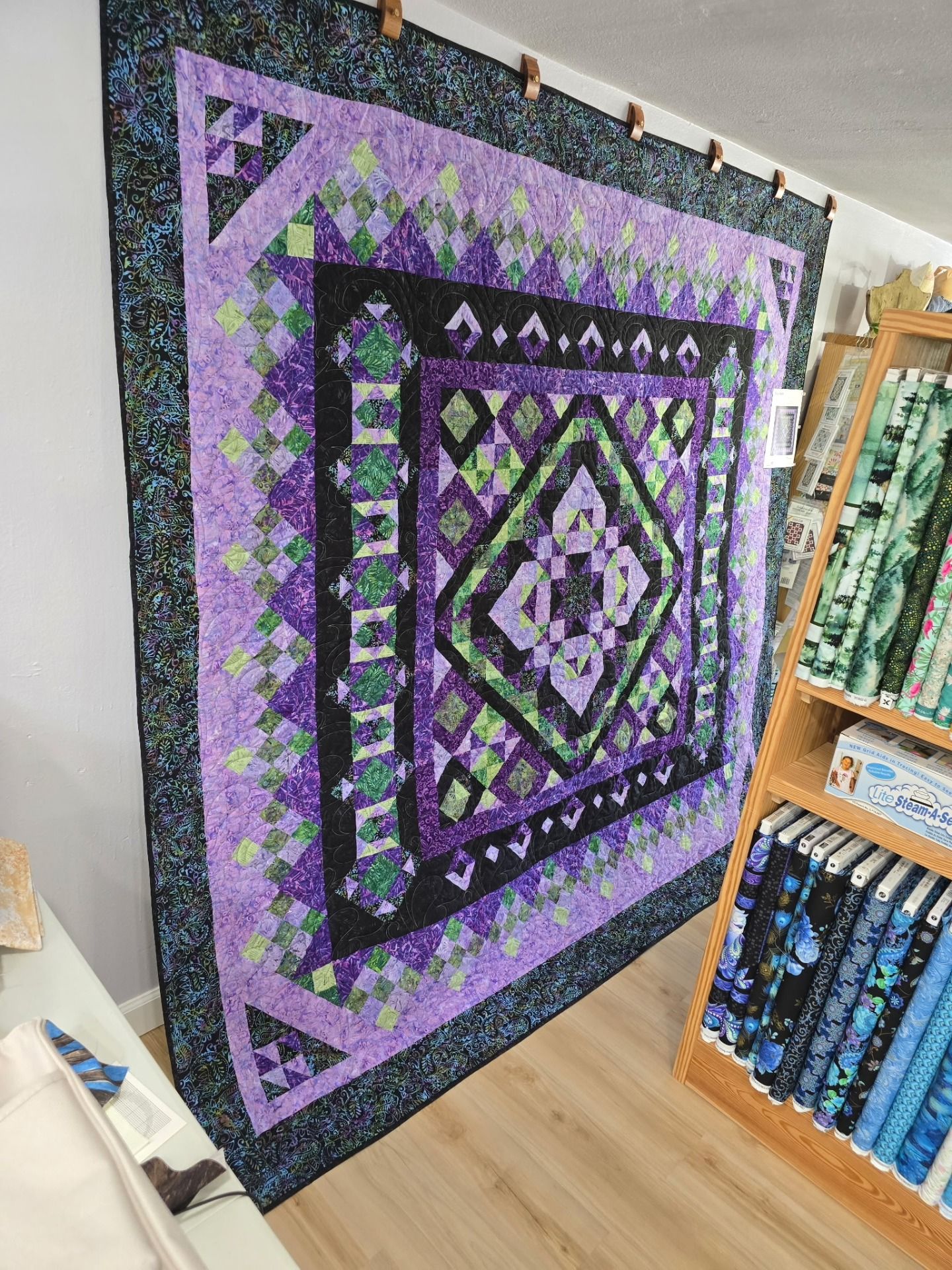 Large purple, black, and green geometric quilt hanging in a fabric shop aisle