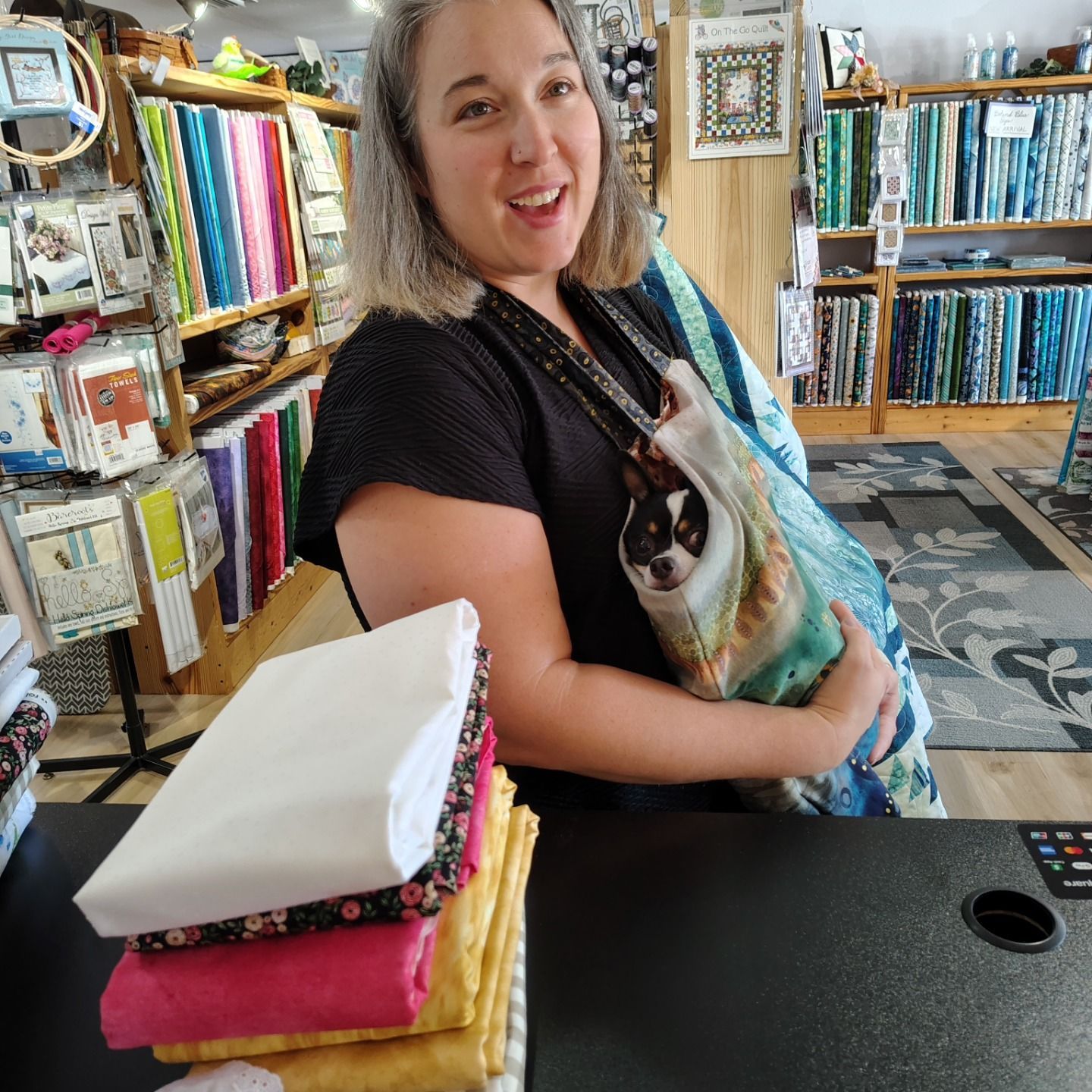 Fabric shop clerk smiling, holding a cat in a sling beside colorful quilt fabrics and folded cloths.