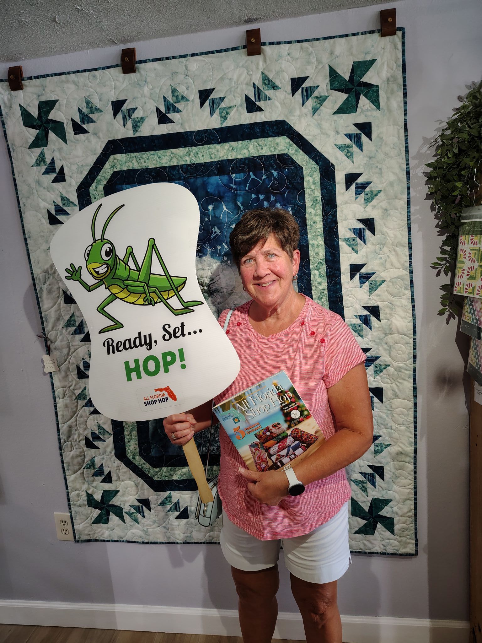 Woman holding a quilt and a round sign that reads “Handy. Set. HOP!” in front of a quilt wall hanging