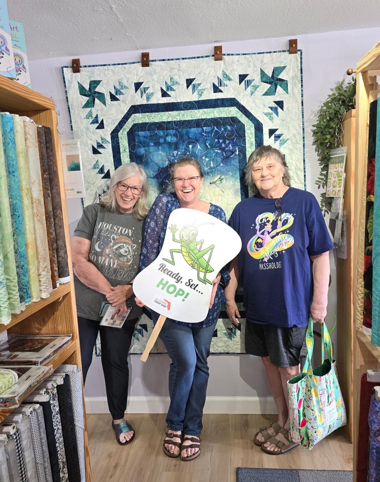 Three smiling people standing in a quilt shop, holding quilts and a tote bag
