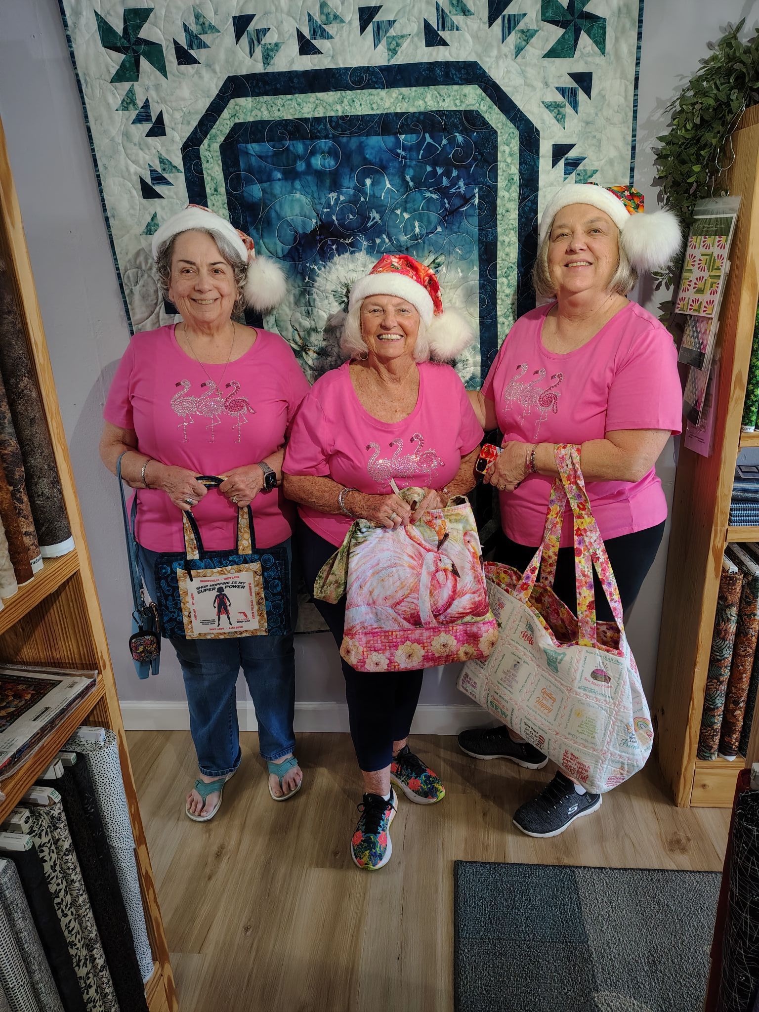 Three women in pink shirts pose with festive bags in front of a holiday quilt display.