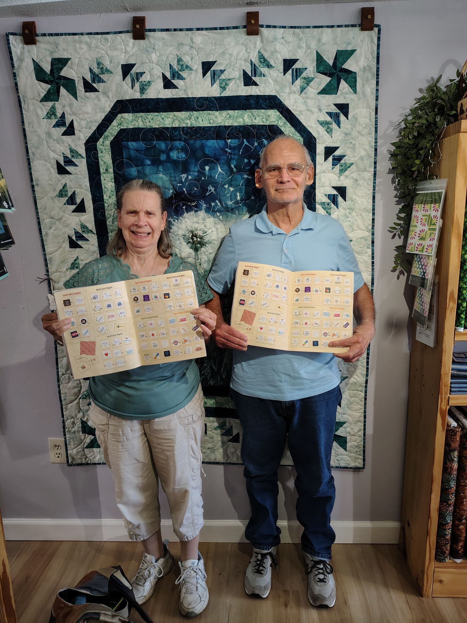 Two people holding quilt blocks in front of a wall-hung quilt display.
