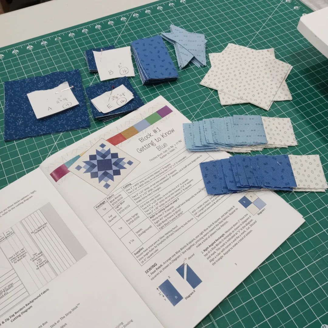 Patchwork fabric pieces and a quilt pattern sheet on a green cutting mat, with blue and white squares arranged nearby