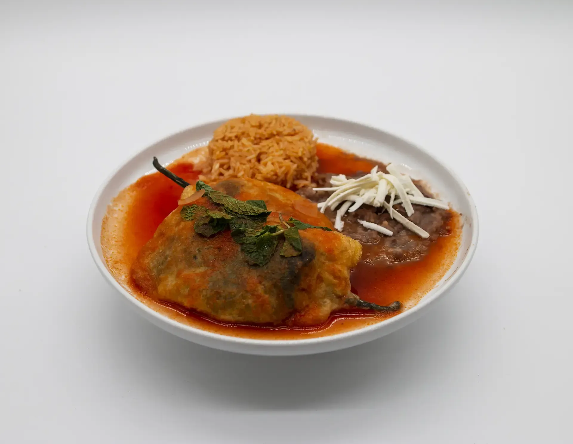 Chili relleno with rice, refried beans, and cheese in red sauce on a white plate.