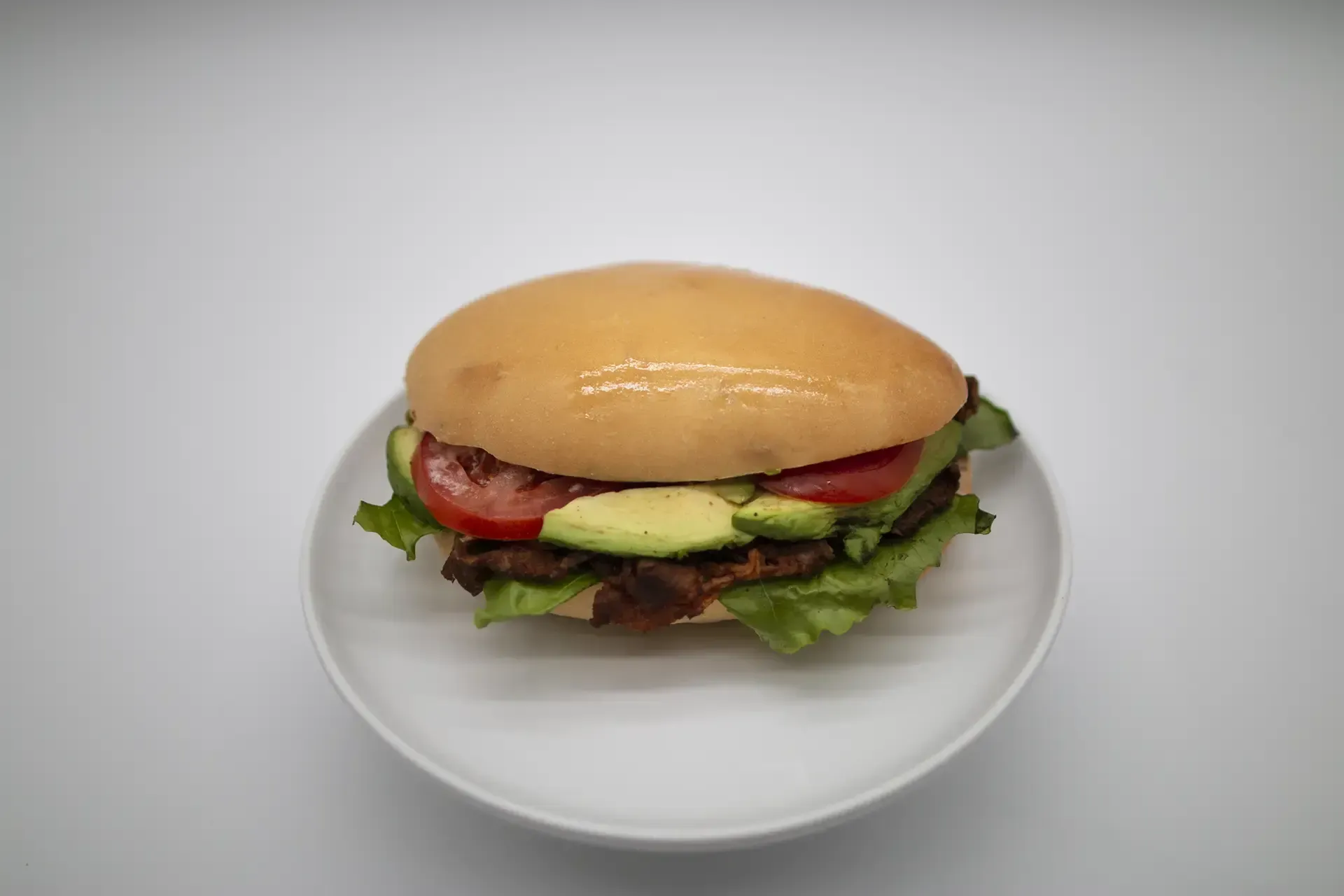 Hamburger with avocado, tomato, and lettuce on a white plate.