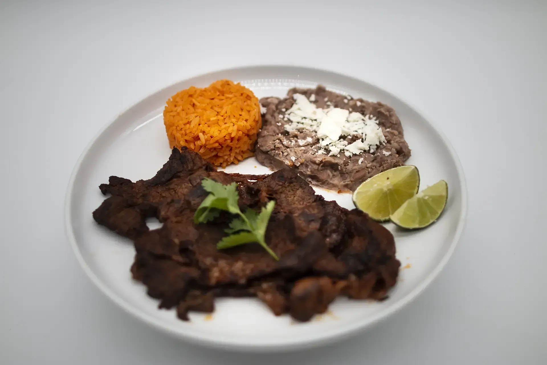 Plate with grilled meat, rice, refried beans, lime wedges, and cilantro.