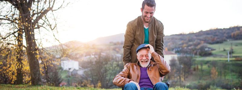 Man in a brown jacket giving a piggyback ride to a smiling person outdoors at sunset
