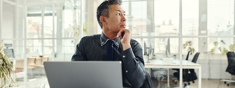 Man at desk in a modern office, gazing thoughtfully out the window, laptop in front of him.