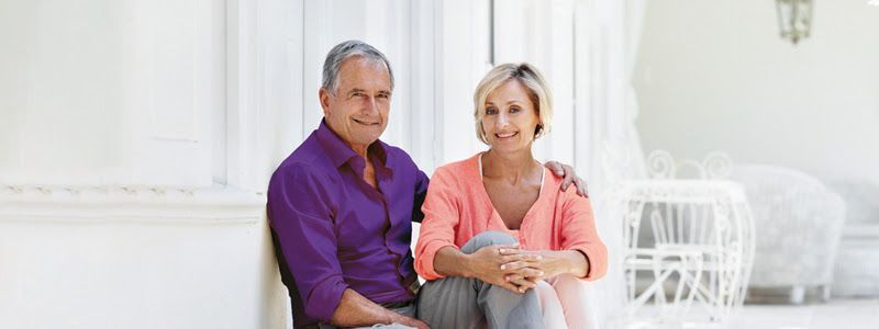 Older couple smiles while sitting on a porch; the man wears purple, the woman wears orange, and white furniture is in the background.
