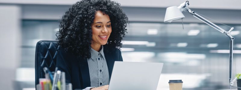 Smiling woman with dark curly hair works on a laptop at her desk in a modern office.