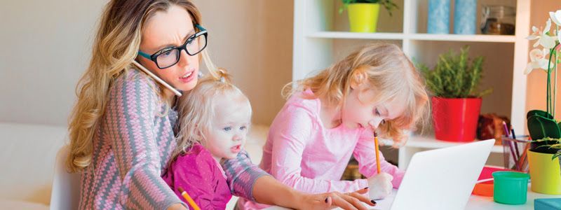 A woman is talking on a cell phone while sitting next to two little girls using a laptop.