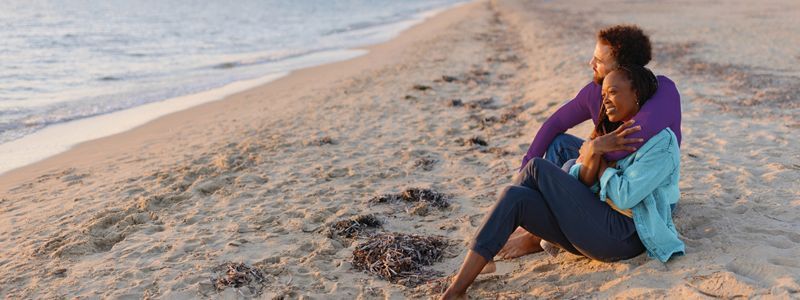 Person sitting on a sandy beach beside the water, wearing a purple top and turquoise shawl