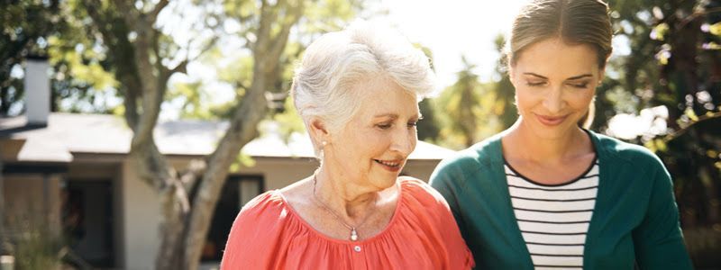 An older and younger woman are walking outside while looking down and smiling.