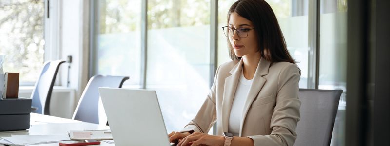 Woman in a beige blazer working on a laptop at a bright office desk by a window