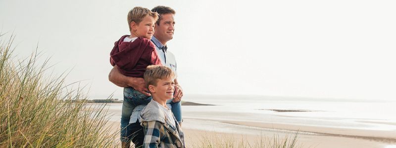 Four people standing on a sandy beach, looking out at the ocean near tall grass.