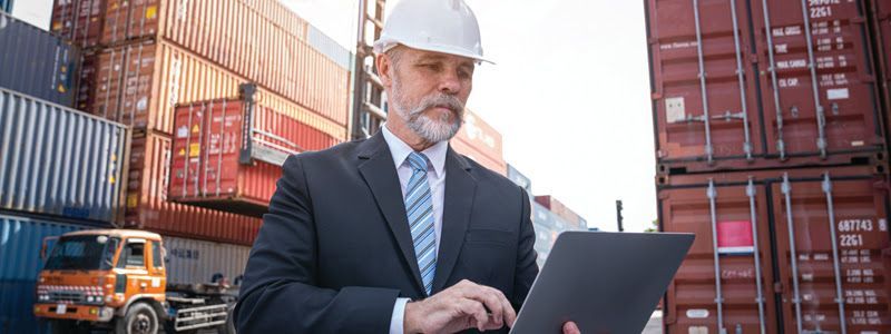 Man in suit and hard hat reviews a document in front of shipping containers in a cargo yard.