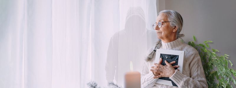 An elderly woman is sitting in front of a window holding a picture frame.