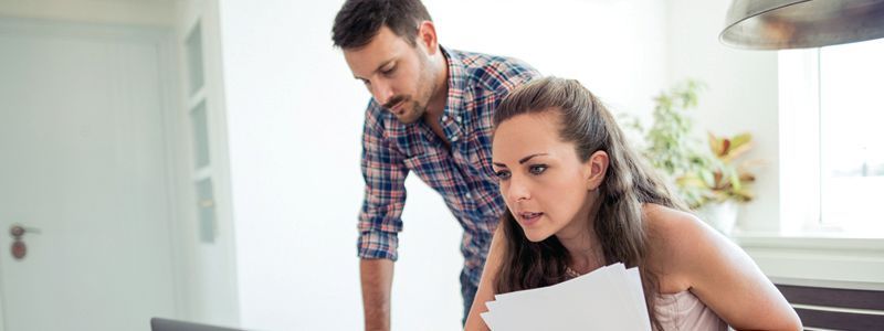 A man and a woman are looking at papers and a laptop in a kitchen.