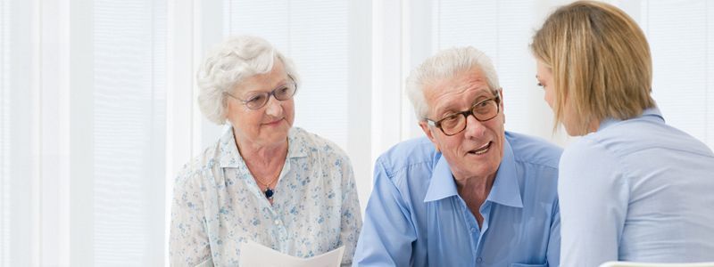 Elderly couple speaking with a younger woman, likely a professional. Light background.
