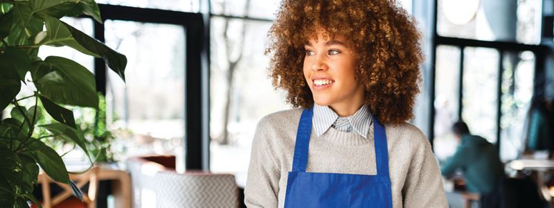Person in a blue apron smiling in a bright workshop or café setting