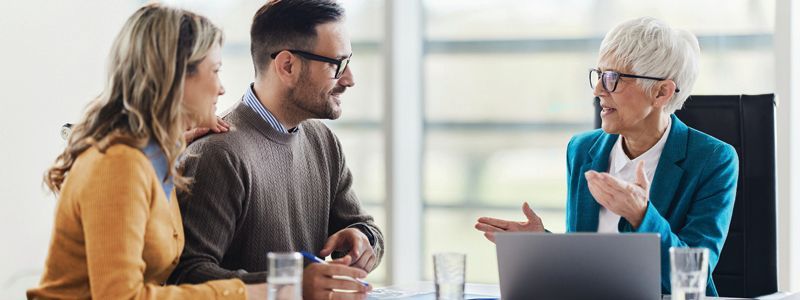 A woman is talking to a man and a woman while sitting at a table with a laptop.