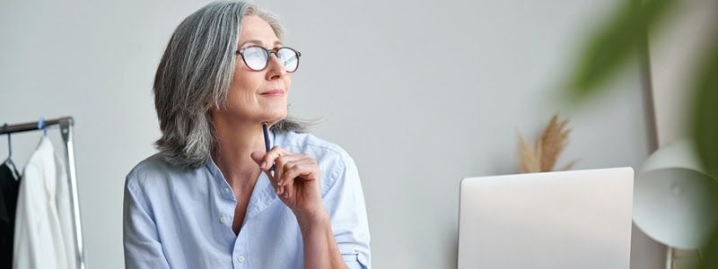 An older woman with glasses thoughtfully looks off to the side, holding a pen.