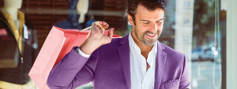 A man in a purple suit is holding shopping bags in front of a store window.
