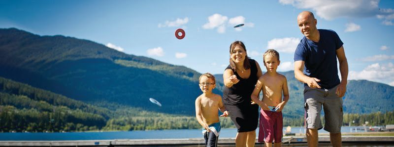 A family is playing a ring toss game by a lake and mountains. Three rings are flying through the air