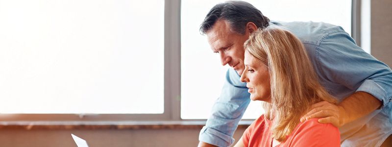 Man and woman looking at a laptop together, woman wearing red and man blue, in front of a bright window.