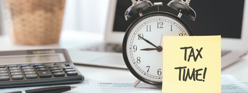 An alarm clock is sitting on a desk next to a calculator and a sticky note that says `` tax time ''.