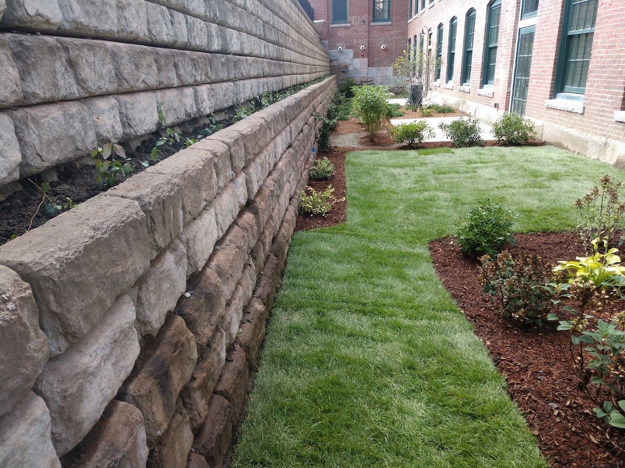 A stone wall surrounds a lush green yard with a brick building in the background.