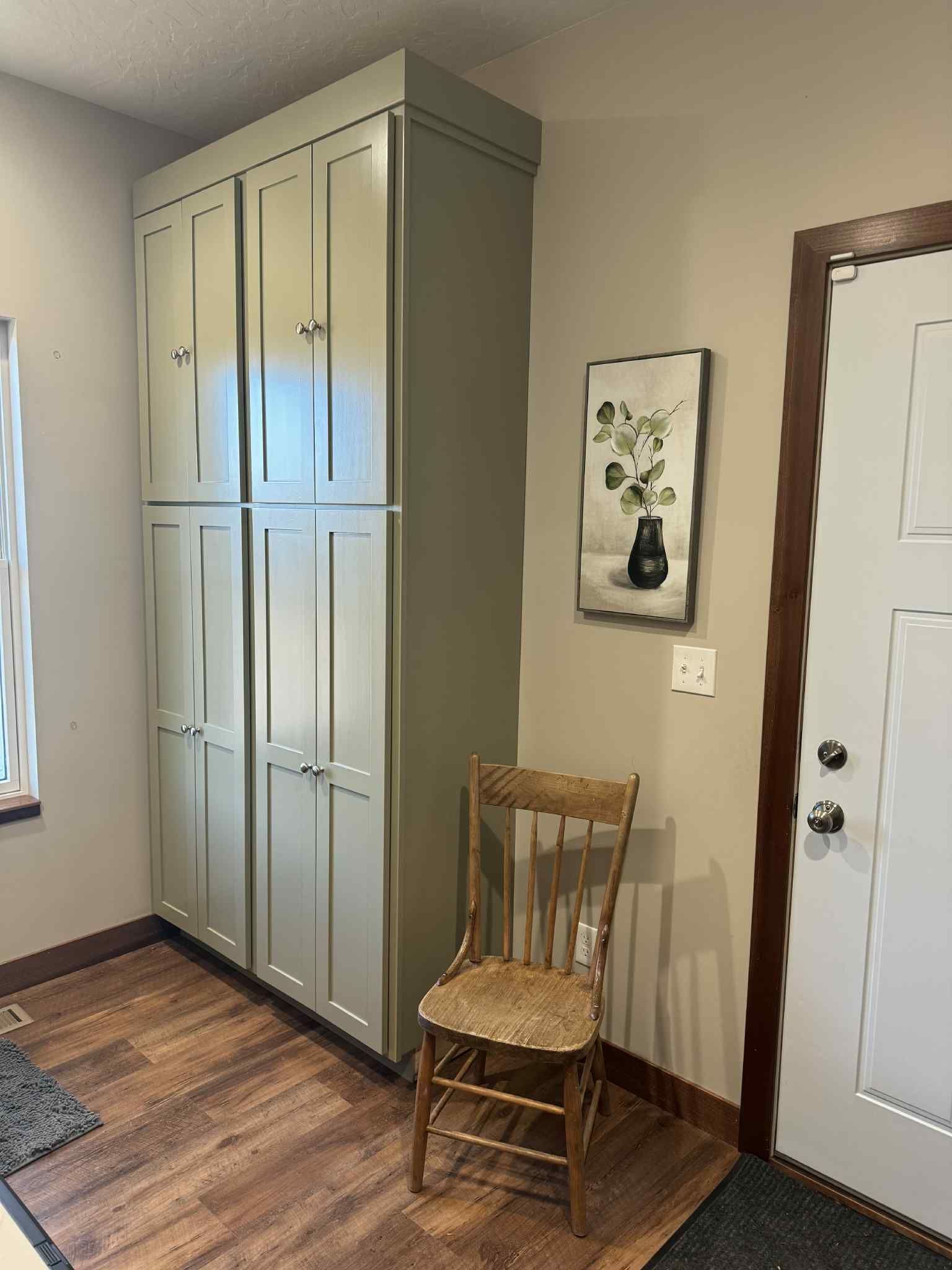 Tall, sage-green cabinets beside a wooden chair and a framed picture, next to a door with brown trim.