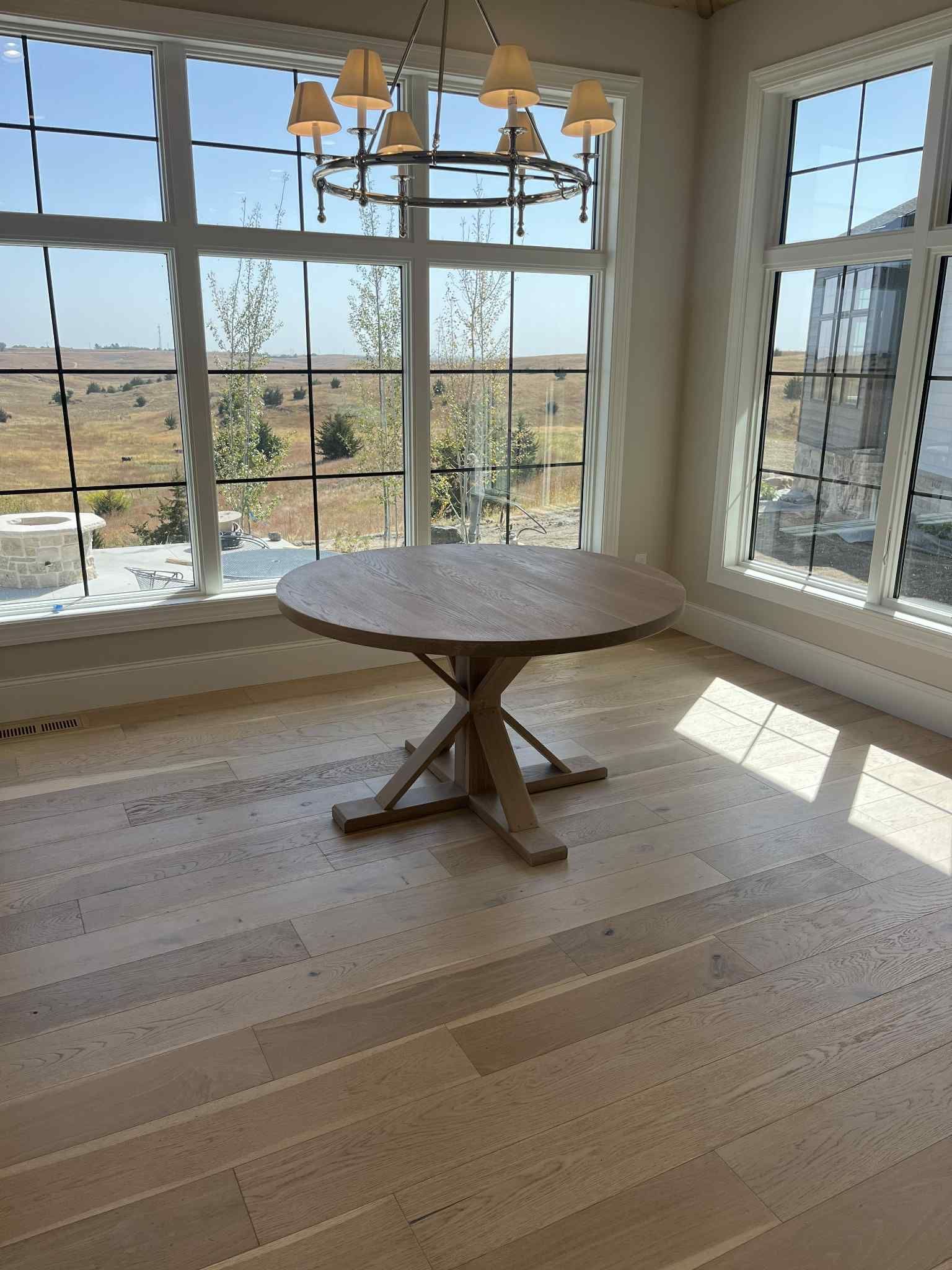 Round wooden dining table in a bright room with large windows, chandelier.