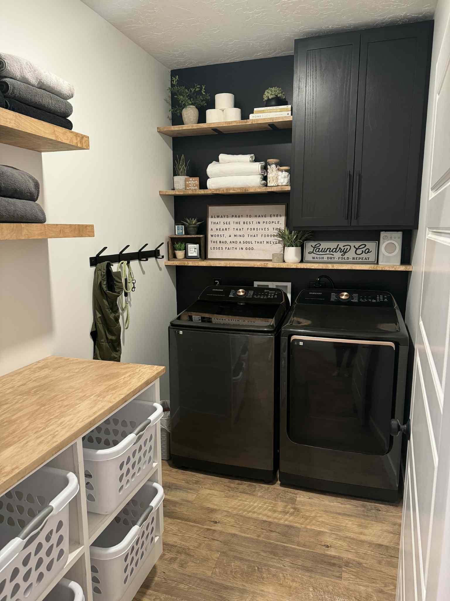 Laundry room with black appliances, black cabinet and accent wall, white and wood shelving, and white baskets.