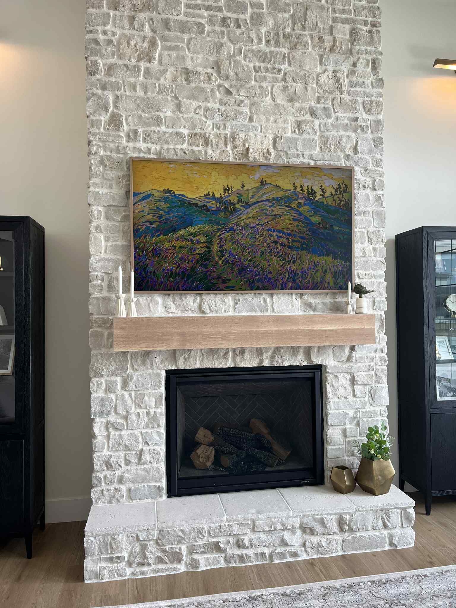 Fireplace with gray brick surround, wood mantel, and a mounted painting; two black cabinets flank the fireplace.